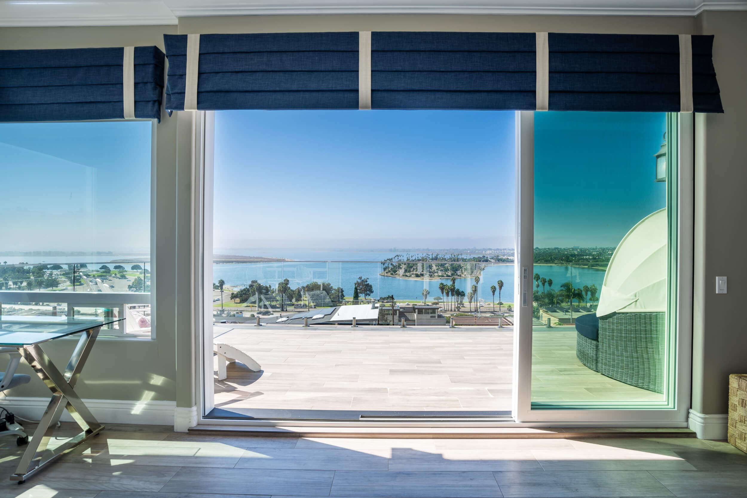 A room with large windows and open sliding door reveals a sunny bay with islands, palm trees, and an outdoor patio. Blue shades.