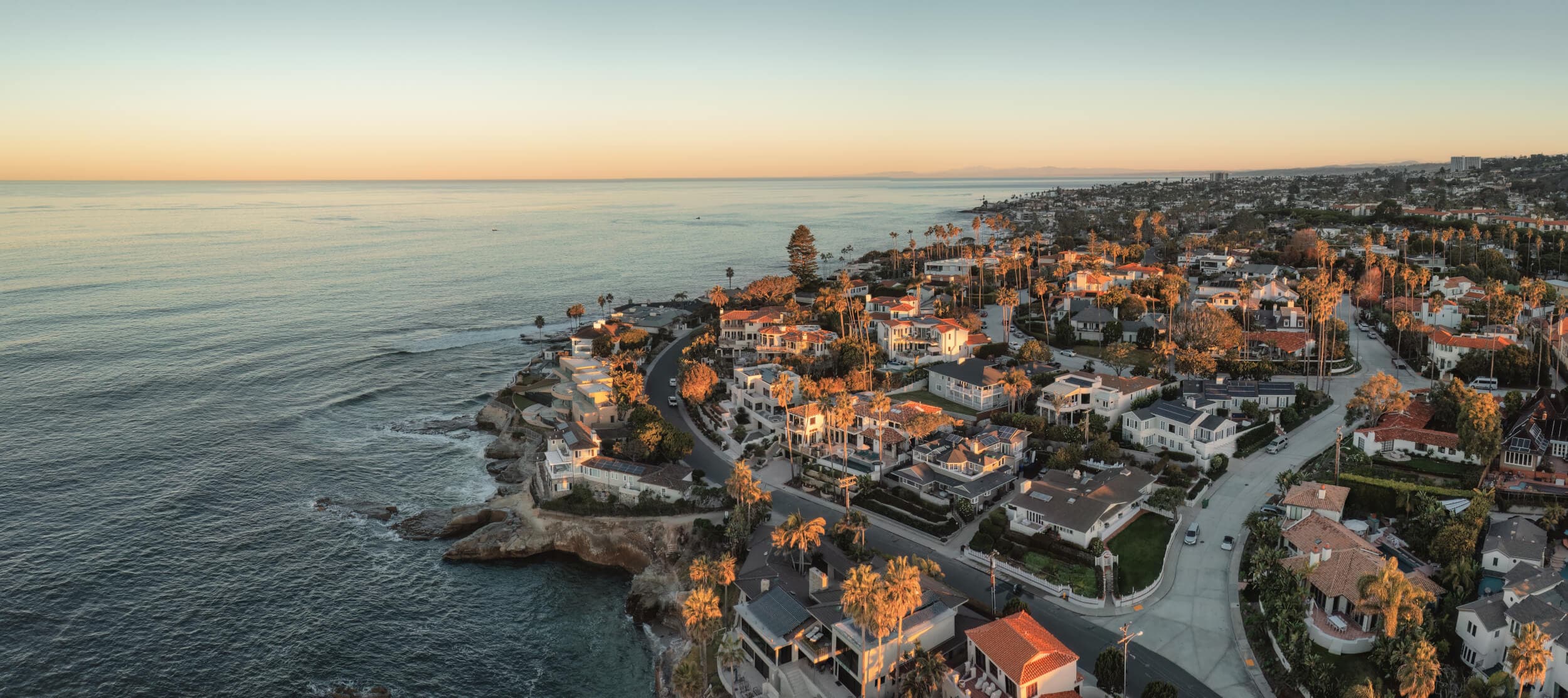 Aerial view of a coastal town at golden hour. The ocean is on the left, with a road winding past houses and palm trees.
